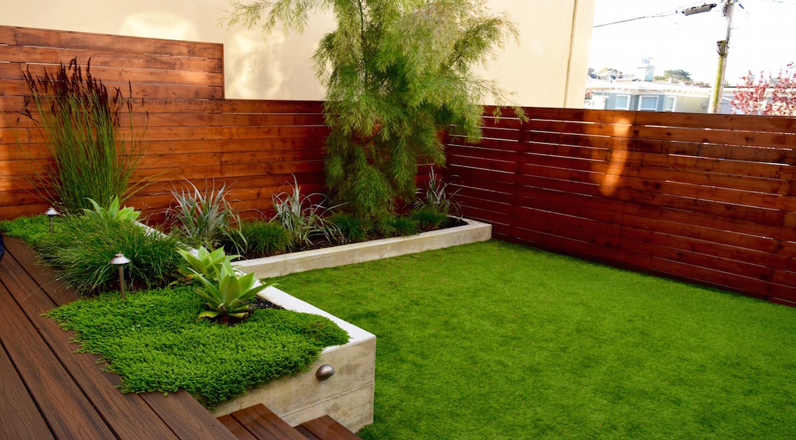 backyard lawn surrounded by concrete planter beds, wooden steps and natural wood fence in the background.
